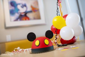 Mickey Mouse cake with ears pictured on a table next to balloons and confetti