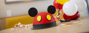a Mickey Mouse cake with ears surrounded by balloons and confetti