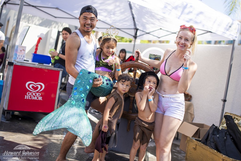 a woman and man pose for a photo with their three children, one of which is dressed like a mermaid, in front of a ship's wheel