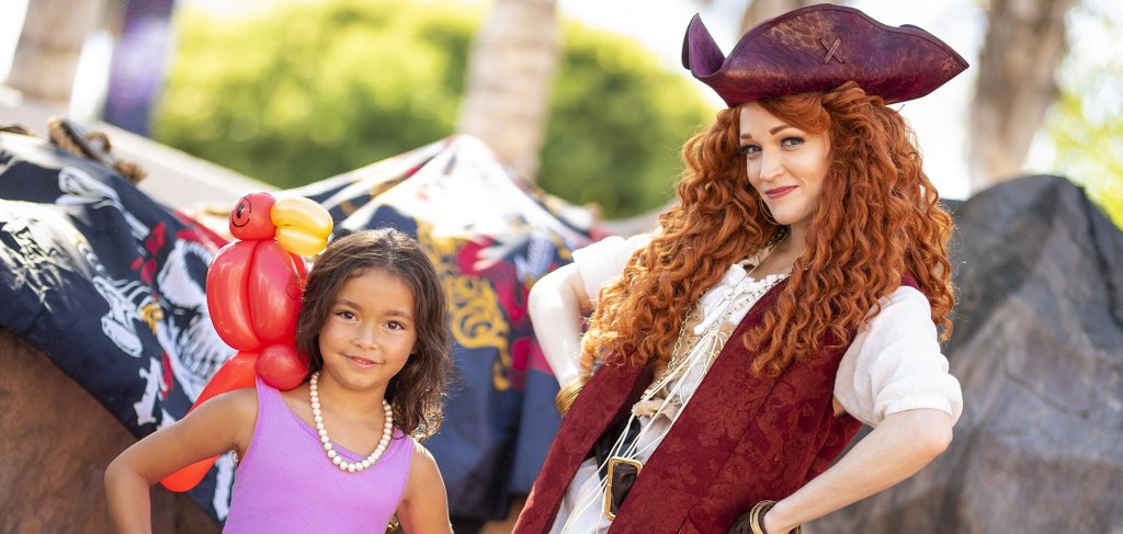 a woman dressed as a pirate poses with a little girl with an inflatable parrot on her shoulder