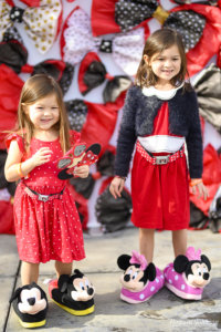 two little girls wear Mickey and Minnie Mouse slippers while smiling in front of a backdrop of polka dot bows