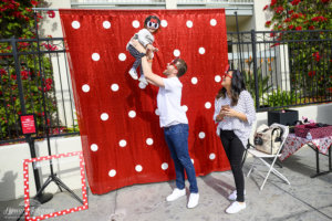 a man holds a little girl in the air in front of a polka dot backdrop while a woman smiles and watches them