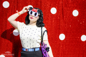 A young woman poses for a picture in front of a polka dot backdrop wearing Minnie Mouse sunglasses