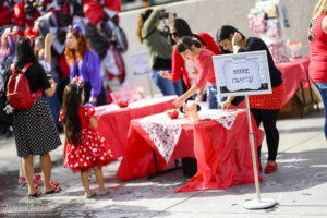 women set up tables labeled from a sign, "Minnie crafts!" as a young girl in a Minnie Mouse polka dot dress watches