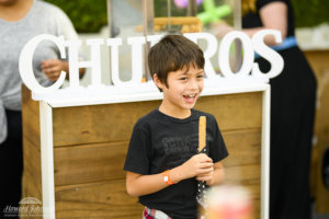 A young boy smiles with a churro in his hand in front of a sign for churros