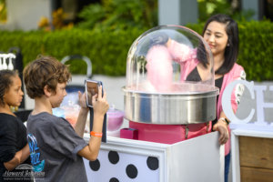 a young woman makes pink cotton candy for two children from a machine while one child takes a photo of it