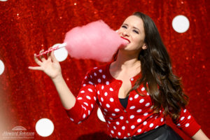 A young woman wearing a polka dot sweater poses in front of a polka dot picture background with pink cotton candy in her mouth