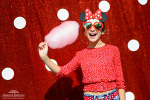 a young girl poses in Disney sunglasses and a polka dot shirt in front of a polka dot picture background with pink cotton candy in her hand