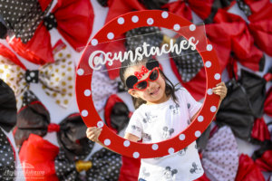 a little girl with Minnie Mouse sunglasses poses for a photo with a circular polka dot frame and social media handle on it
