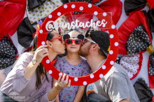 a mother and father give their daughter a kiss on the cheek while posing for a photo with a circular polka dot frame and social media handle