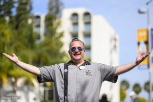 A man wearing sunglasses and a Disneyland shirt holds his hands in the air and smiles, pictured in front of the Howard Johnson Anaheim hotel